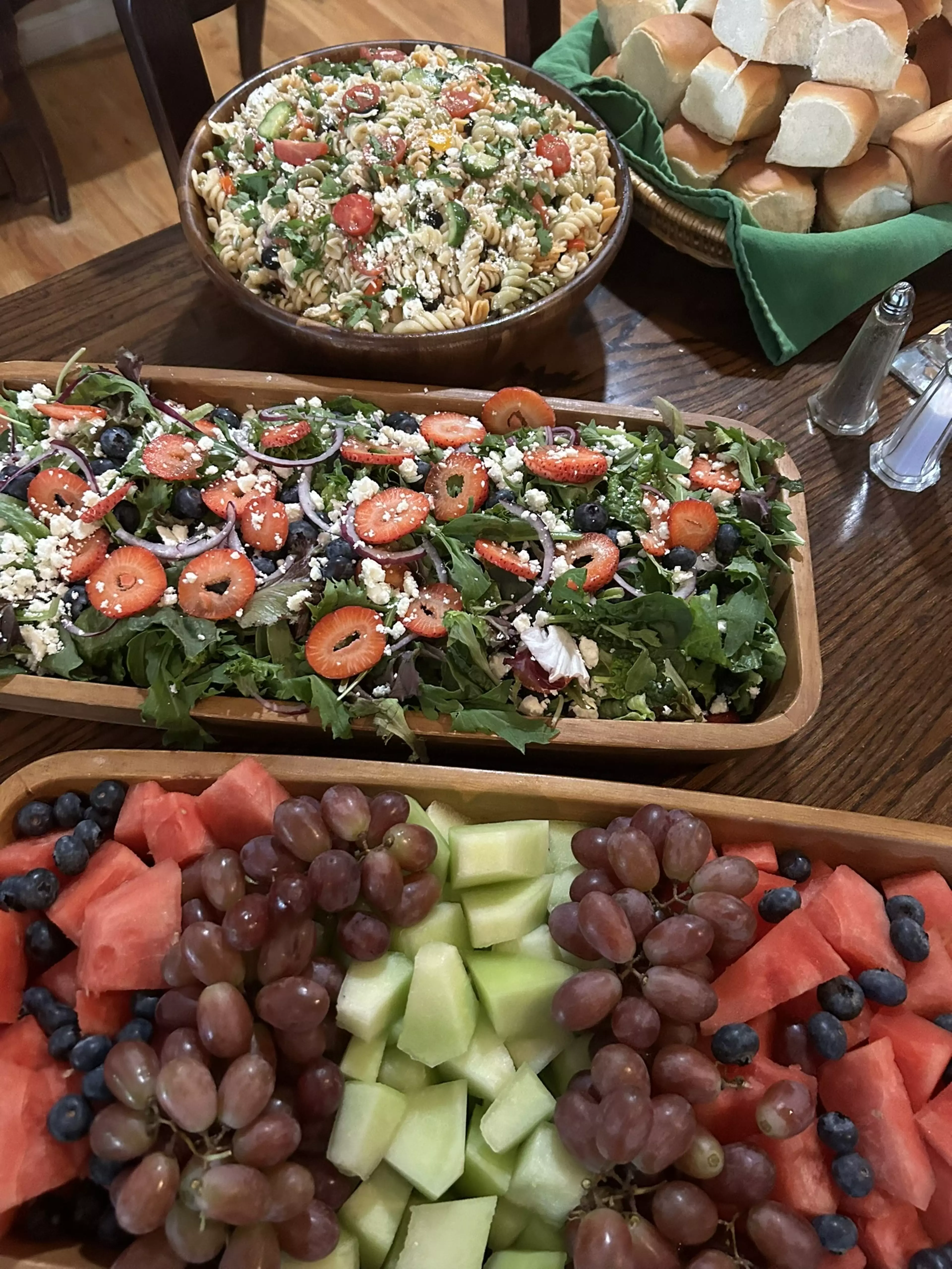 Bowls full of salad, pasta salad, bread, and fruits set out on a wooden table.