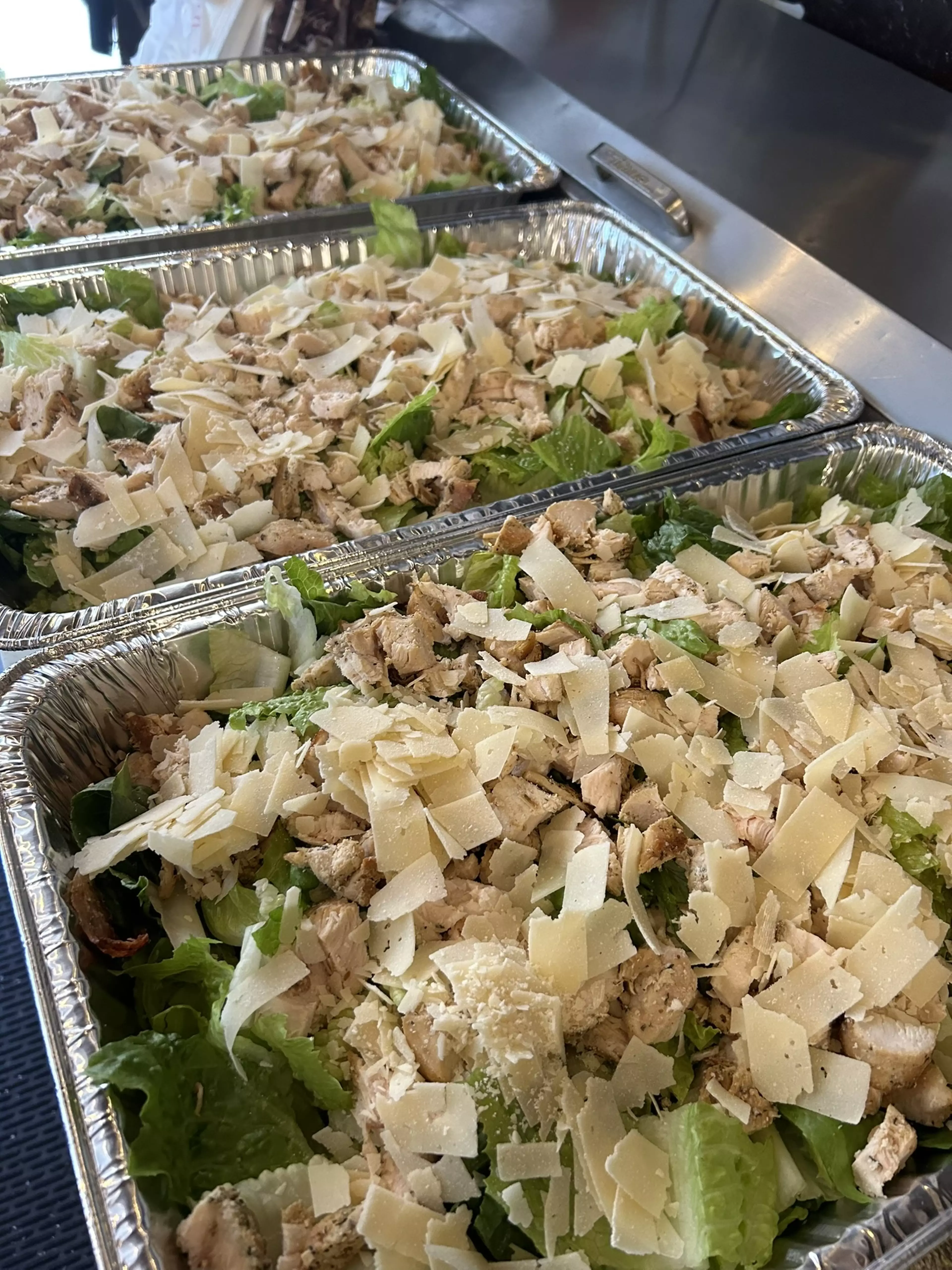 Several trays of chicken cesar salad arranged on a metal countertop.