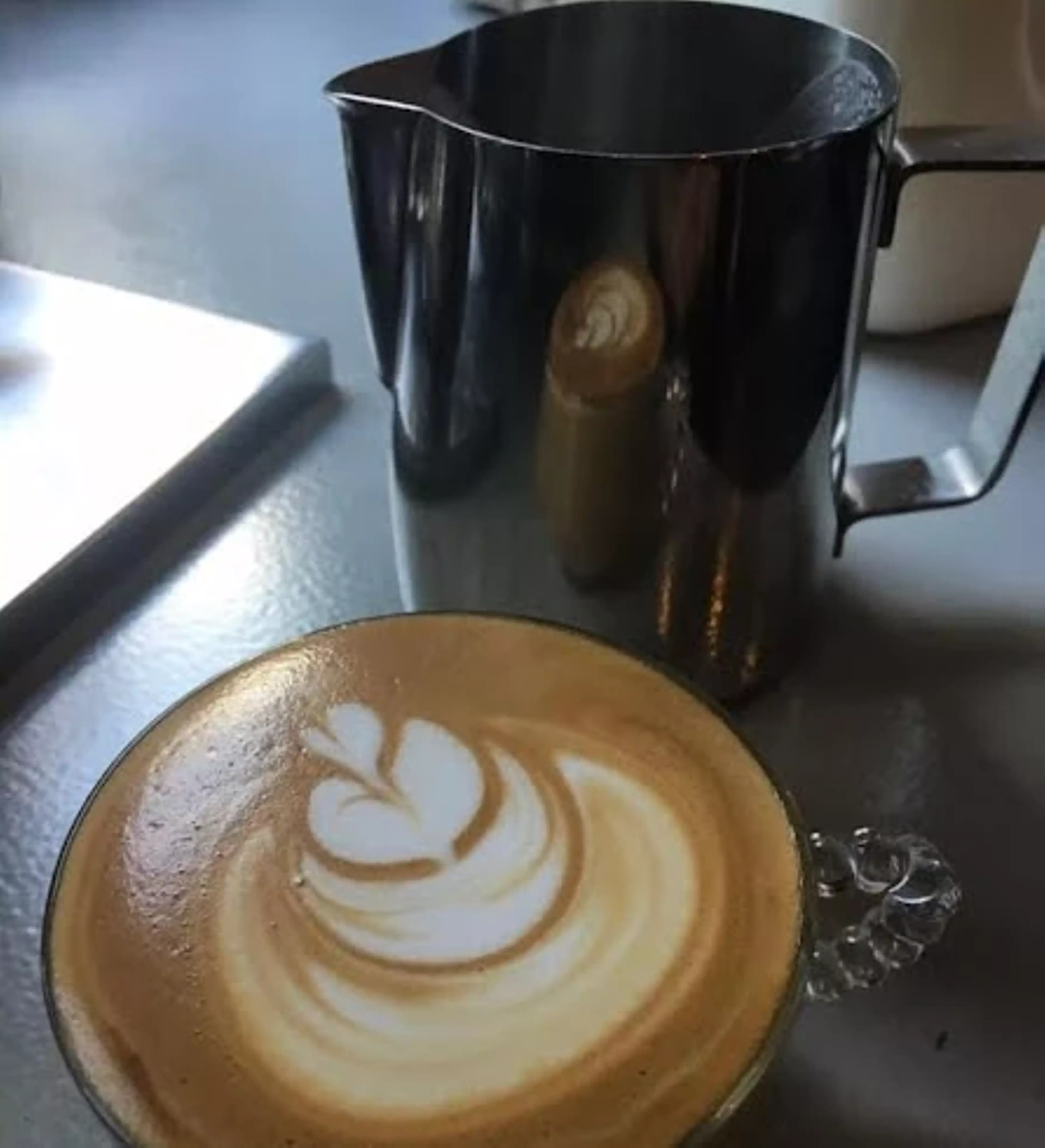 An elaborate latte with foam art sits in front of a metal pitcher.